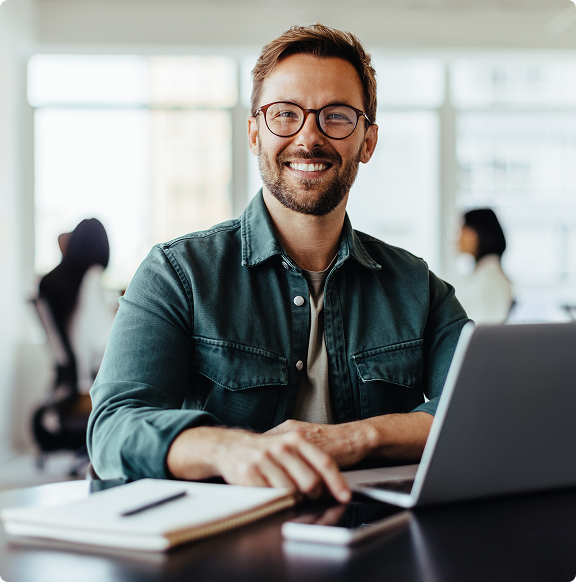 homem em um escritório sentado em uma mesa com computador à sua frente, ele está olhando diretamente para a câmera e sorrindo. 
