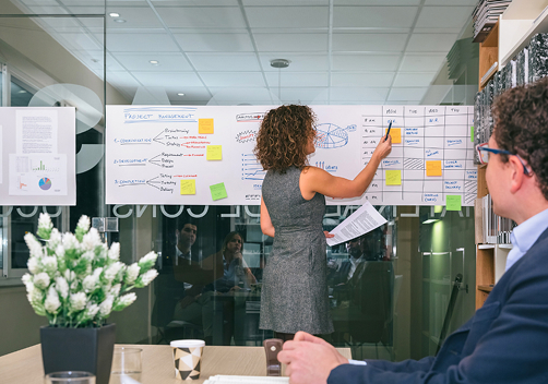 sala de reuniões de uma empresa com papéis colados na parede enquanto duas pessoas discutem os planos e projetos